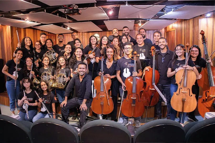 Grupo reunido em sala de ensaio, músicos com violoncelos e clarinetes, camisetas pretas.