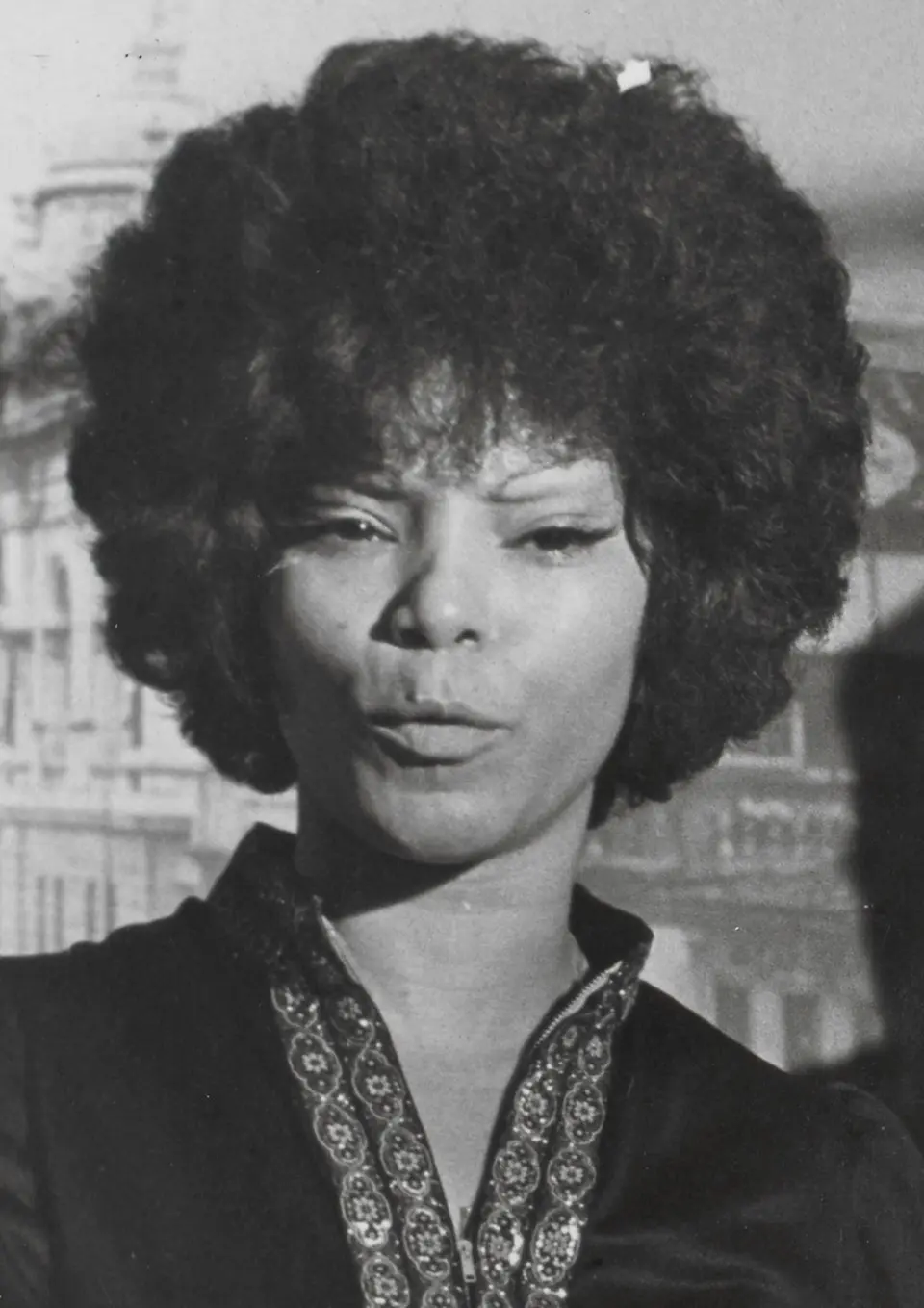 Uma foto preto e branco mostra uma pessoa com cabelos afro olhando para baixo, usando um top decorativo.