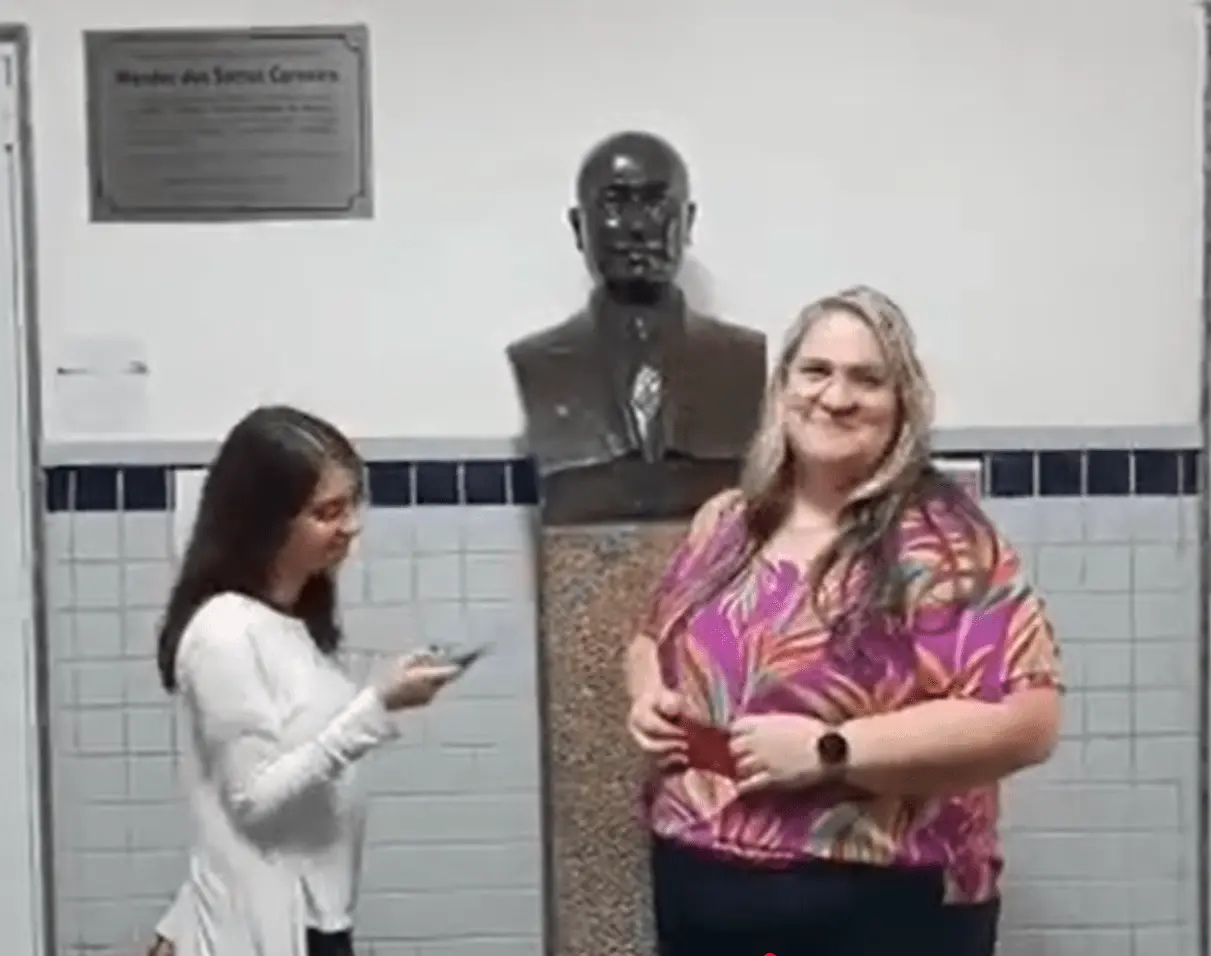 Duas mulheres posam junto a busto de bronze sobre pedestal, em ambiente interno com parede de azulejos.