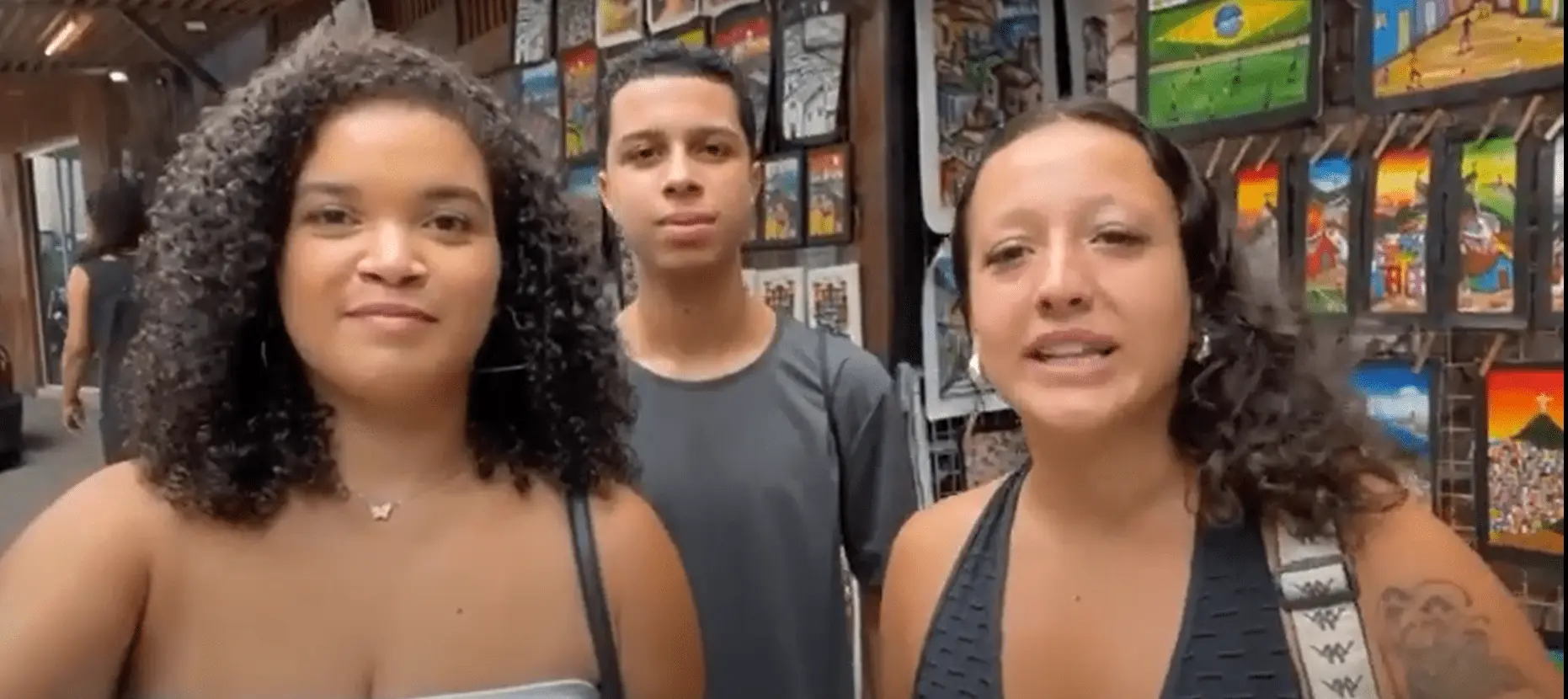 Três pessoas em loja, duas mulheres e um homem, com parede colorida de quadros atrás.