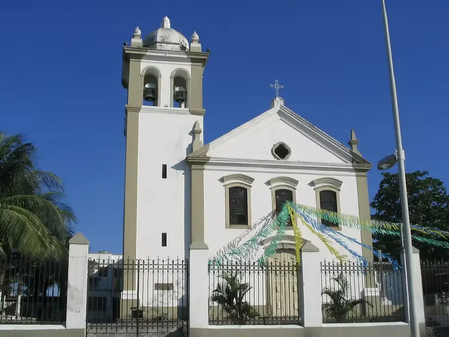 Igreja branca com cúpula e torre, grade preta, árvore verde, céu azul claro.