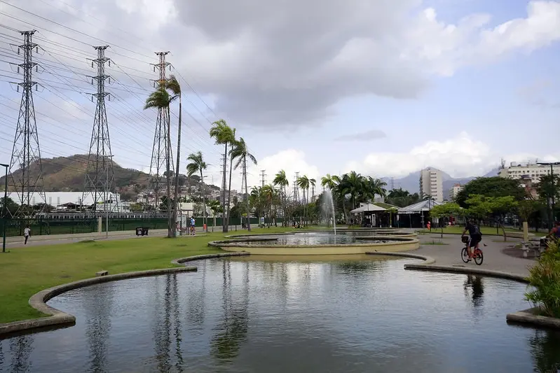 Parque com torres de transmissão, palmeiras, chafariz, pessoas caminhando e bicicleta, edifícios e montanha.