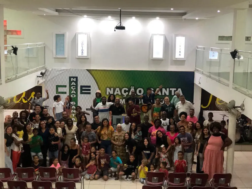 Grupo de pessoas em igreja, com banner "Nação Santa" e placa "CENAÇAO SANTA".