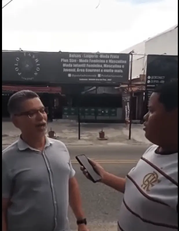 Dois indivíduos em ambiente urbano, homem olhando para mulher segurando celular.
