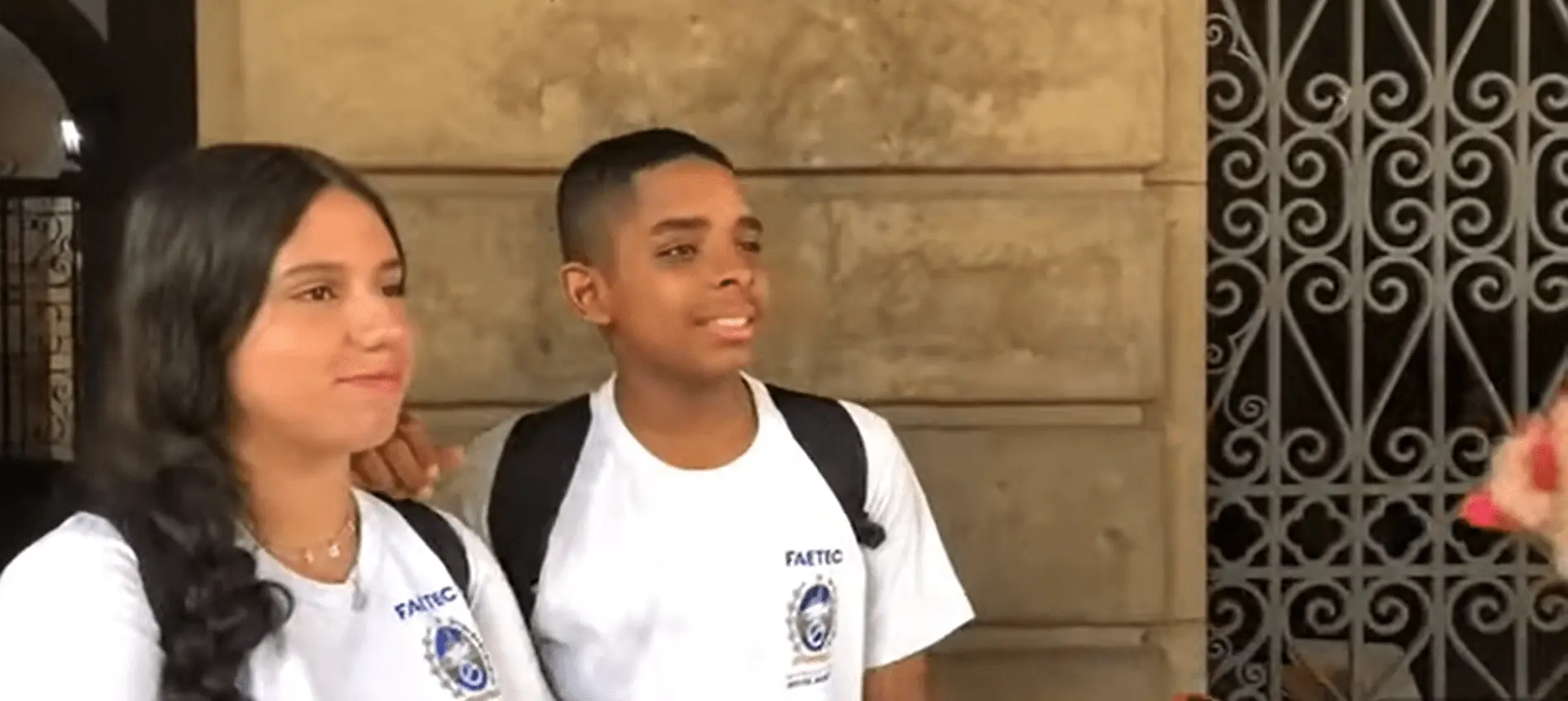 Dois estudantes sorridentes em frente a um edifício de pedra com grades metálicas.
