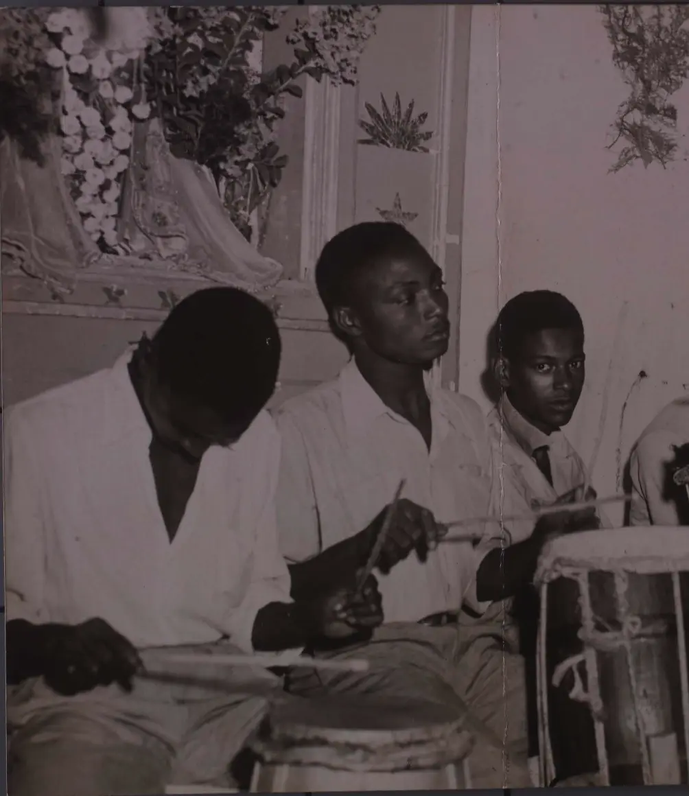 Três homens sentados lado a lado, tocando instrumentos musicais, com decorações florais na parede acima.