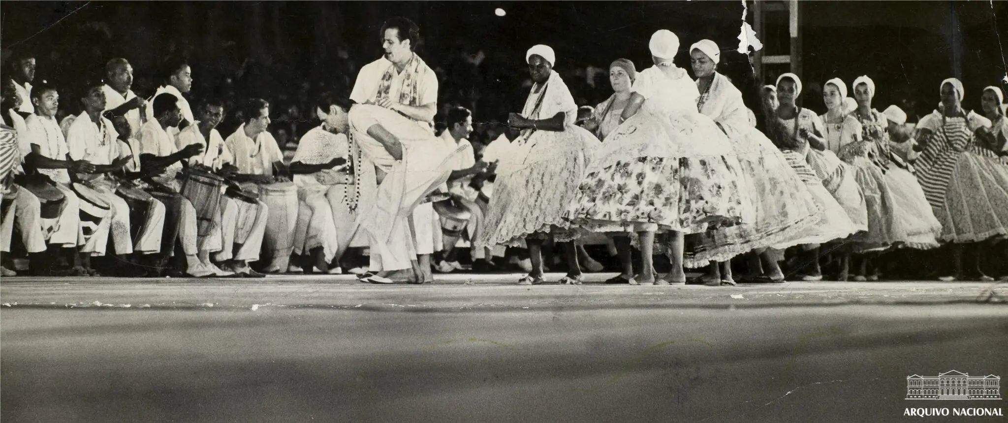 Fotografia preto e branco de evento cultural com músicos tocando instrumentos tradicionais e dançarinos celebrando.