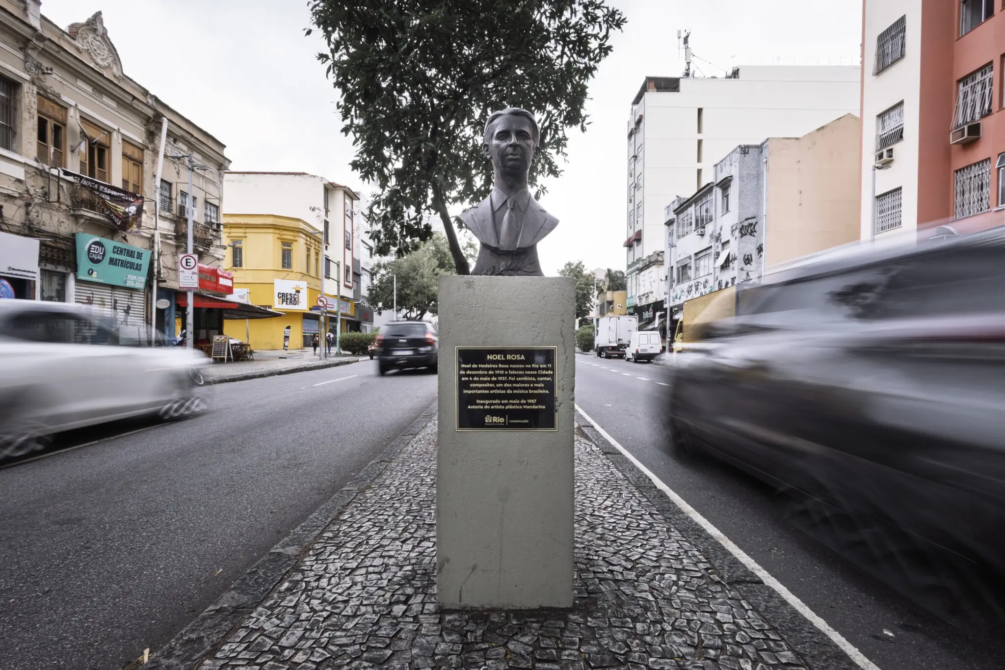 Estátua de Noel Rosa em meio-fio urbano, com edifícios antigos à esquerda e modernos à direita.