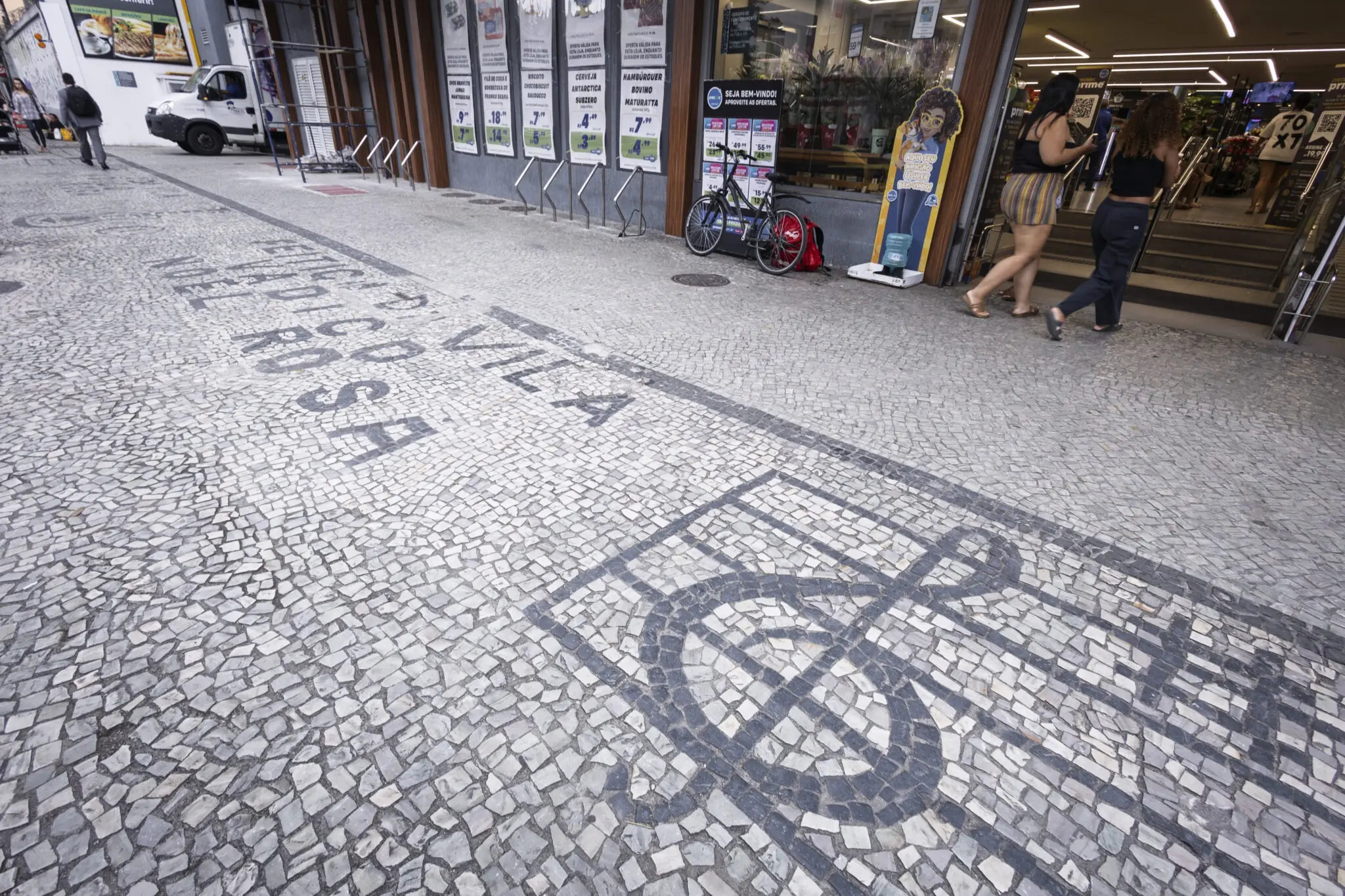 Calçada com mosaico artístico, lojas e pessoas caminhando.