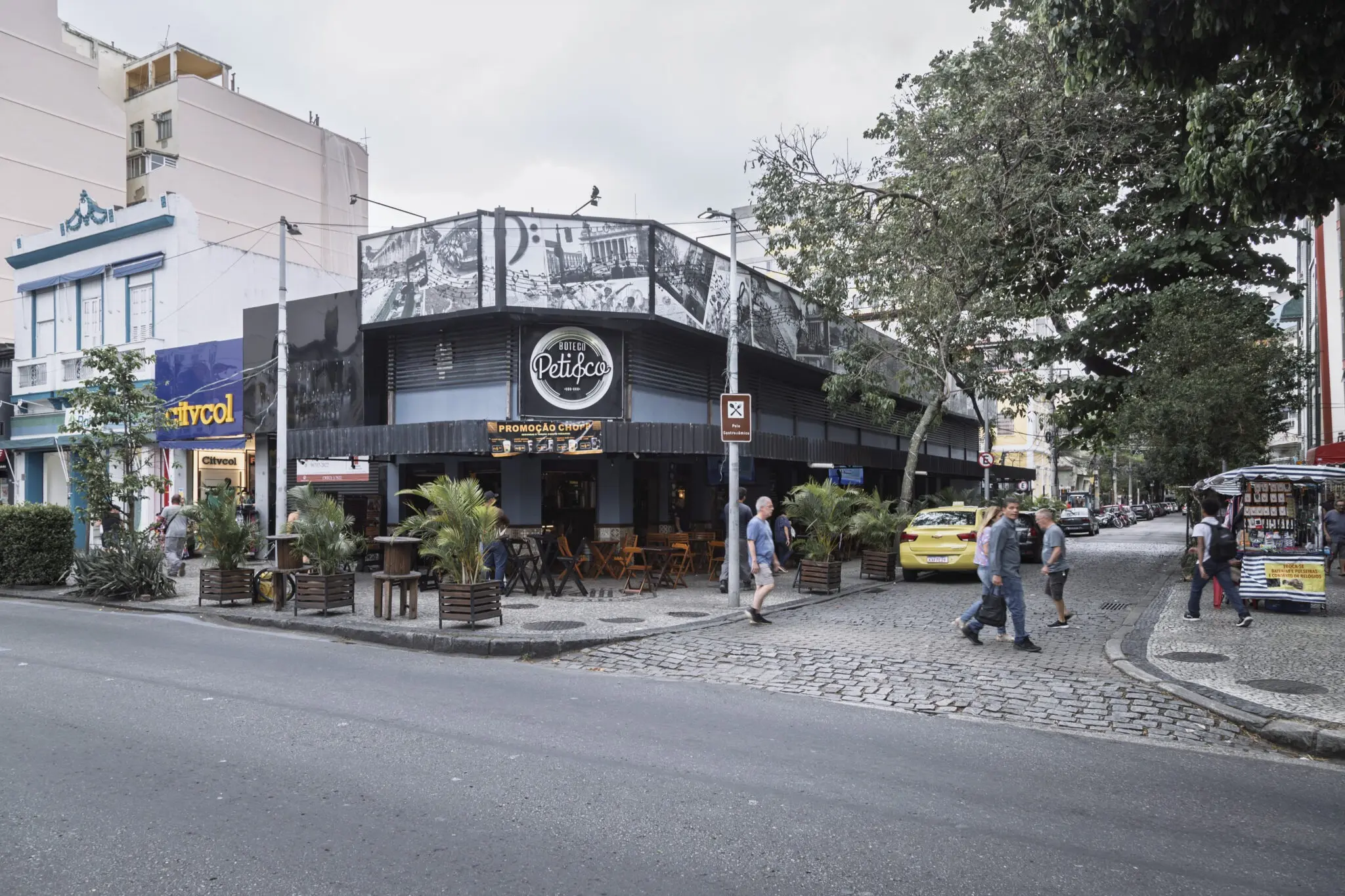 Café "Petisco da Vila" em esquina urbana com edifícios e árvores.