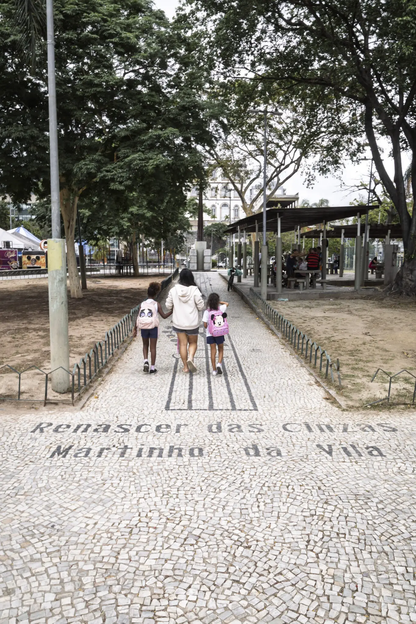 Praça com pessoas caminhando, árvores e edifícios ao fundo.