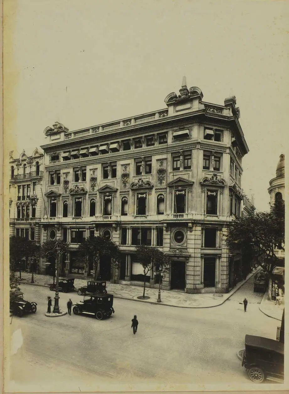 Edifício clássico em esquina com árvores e carros antigos.