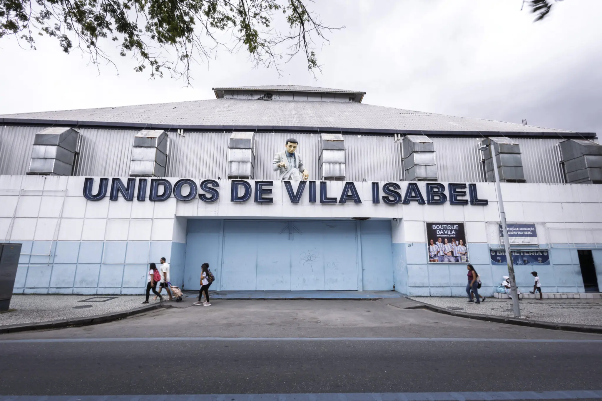 Prédio azul com inscrição "Unidos de Vila Isabel", pessoas em samba e caminhando.