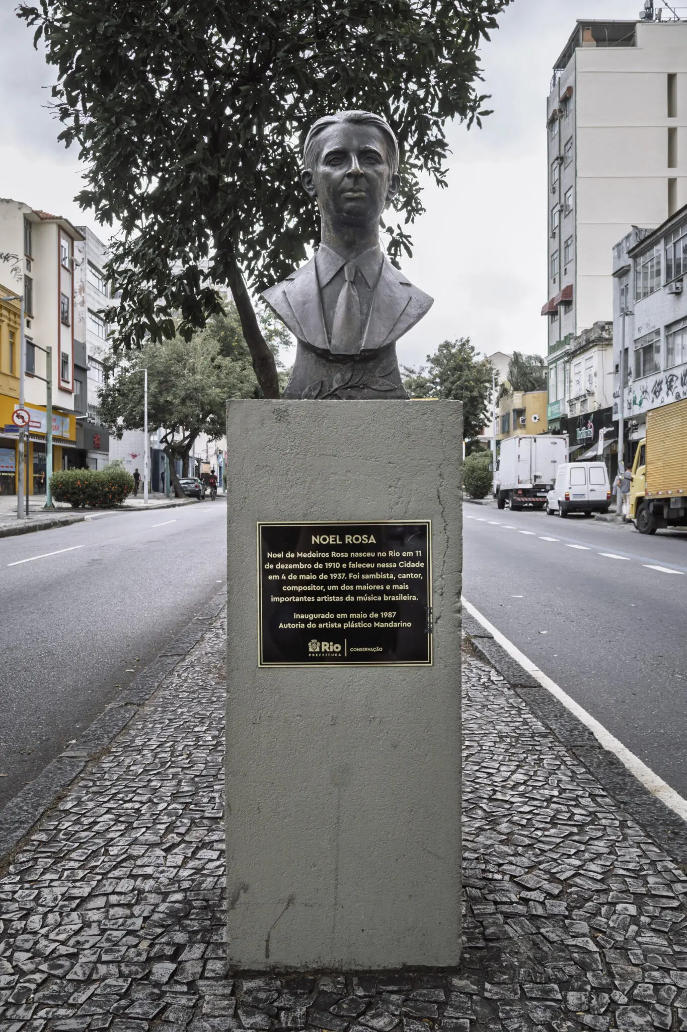 Monumento com estátua de Noel Rosa em praça, árvore verde e edifícios ao fundo.