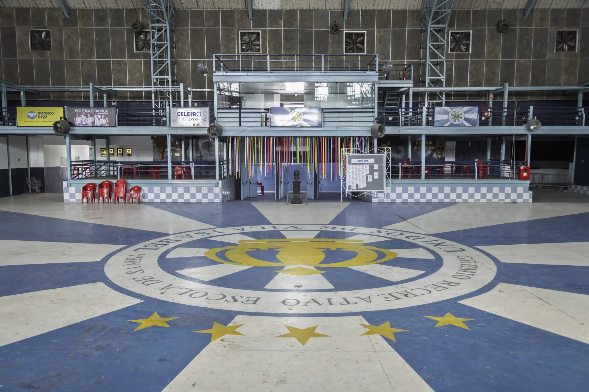 Quadra esportiva com piso azul e branco, cadeiras vermelhas, logotipo com estrelas amarelas e coroa dourada.