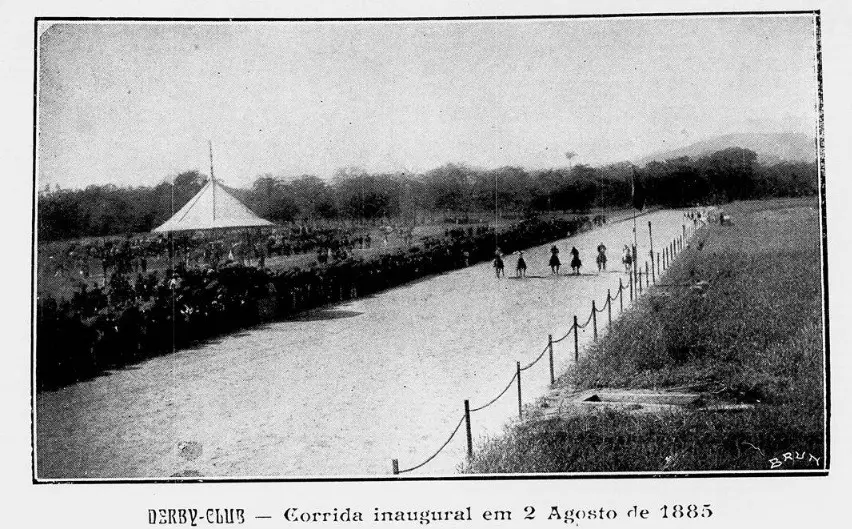 Campo de corrida com cavalos em movimento, tenda branca à esquerda, cerca de cordas ao longo do lado direito, árvores e colina no fundo.