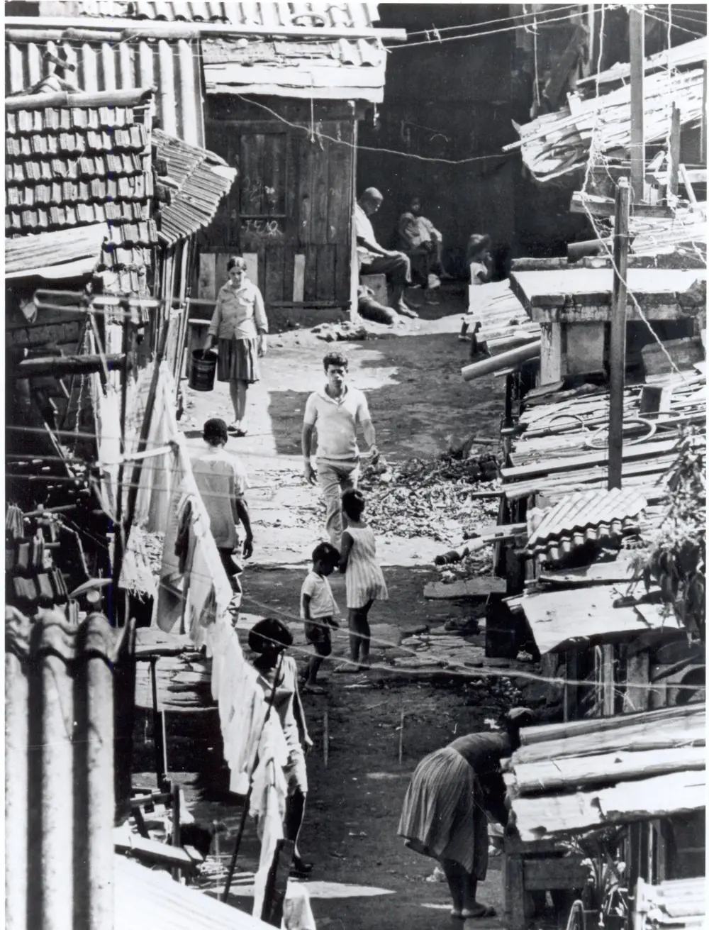 Fotografia preto e branco de favela com barracos e pessoas caminhando.