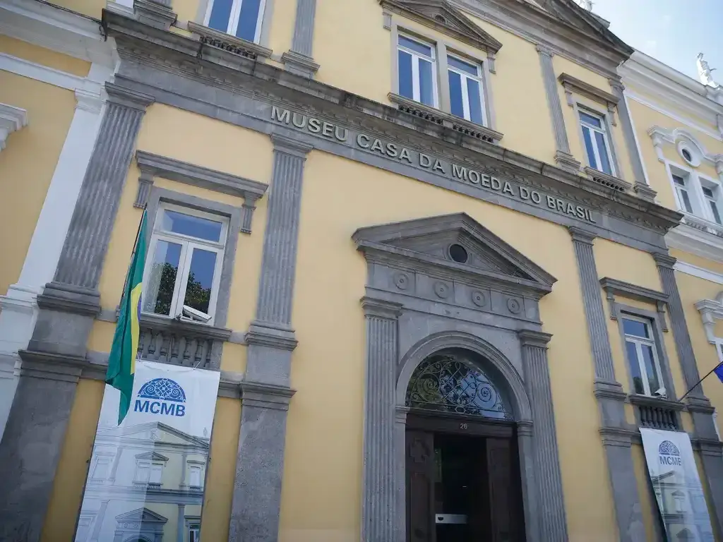 Edifício amarelo com bandeira verde e amarela, banners azuis e entrada marrom.