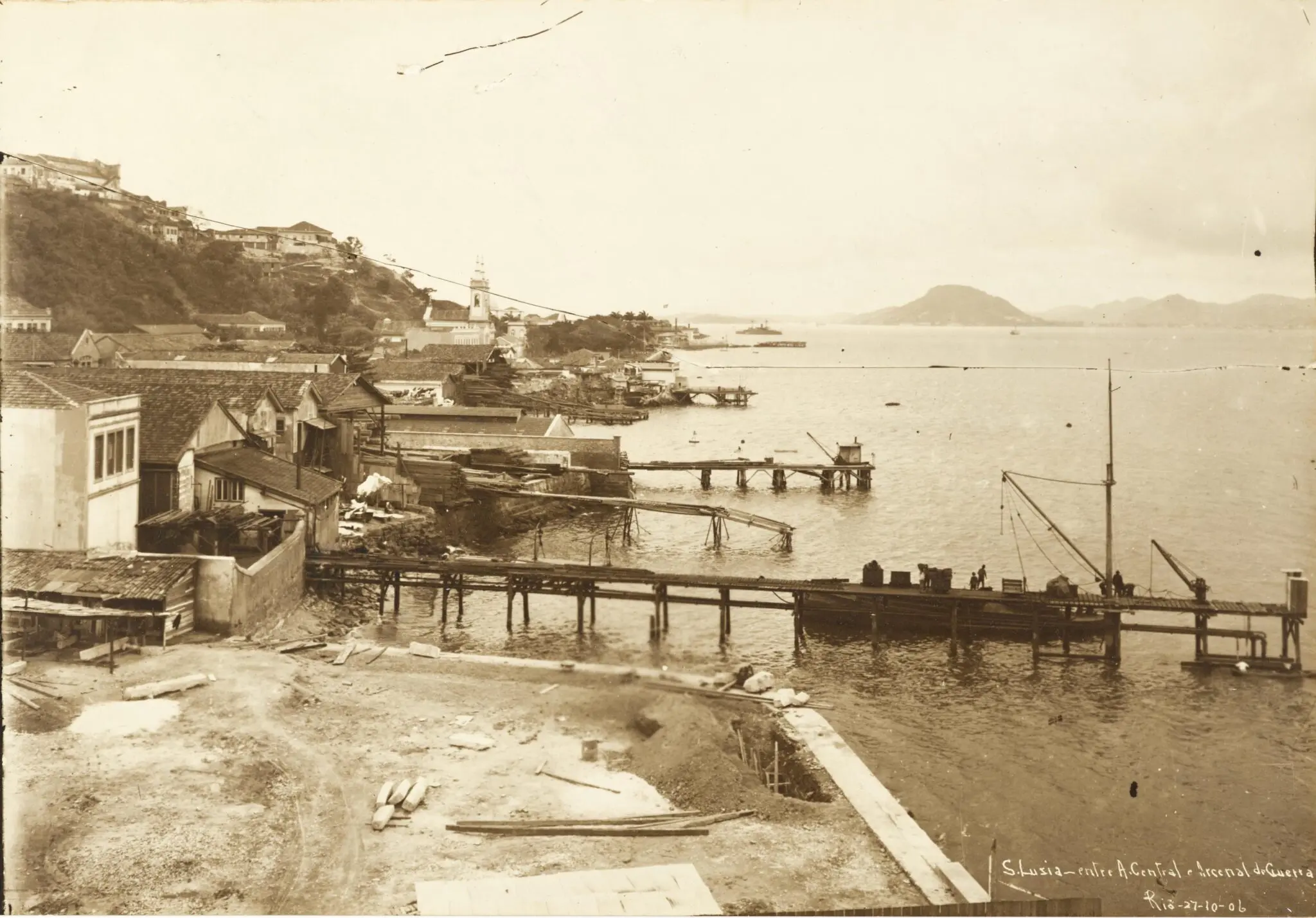 Praia costeira com casas, porto e colina, dia ensolarado.