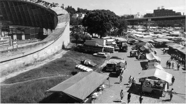 Feira local com barracas e pessoas caminhando, em preto e branco.