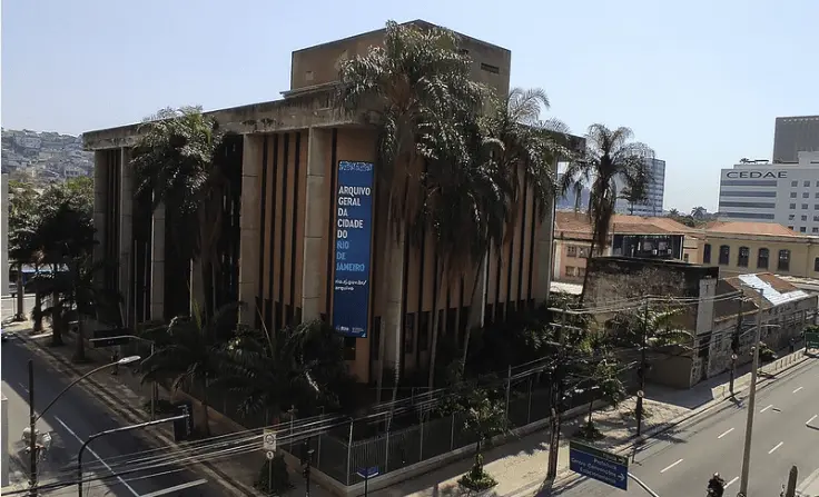 Edifício moderno em área urbana com palmeiras e construções antigas ao fundo.