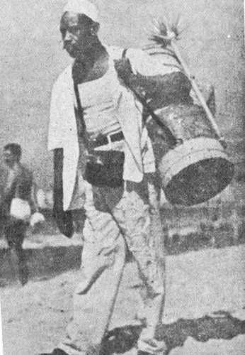 Homem caminhando com recipiente de metal, chapéu branco e camisa branca, fundo com outra pessoa de costas.