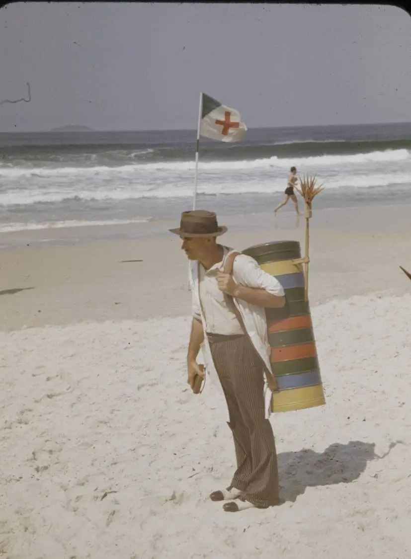 Homem caminhando na praia com cilindro colorido, bandeira da Cruz Vermelha ao fundo.