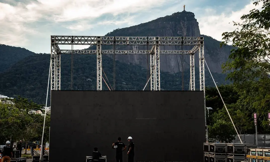 Cenário montado para evento ao ar livre com montanha, estátua, estrutura metálica e pessoas.