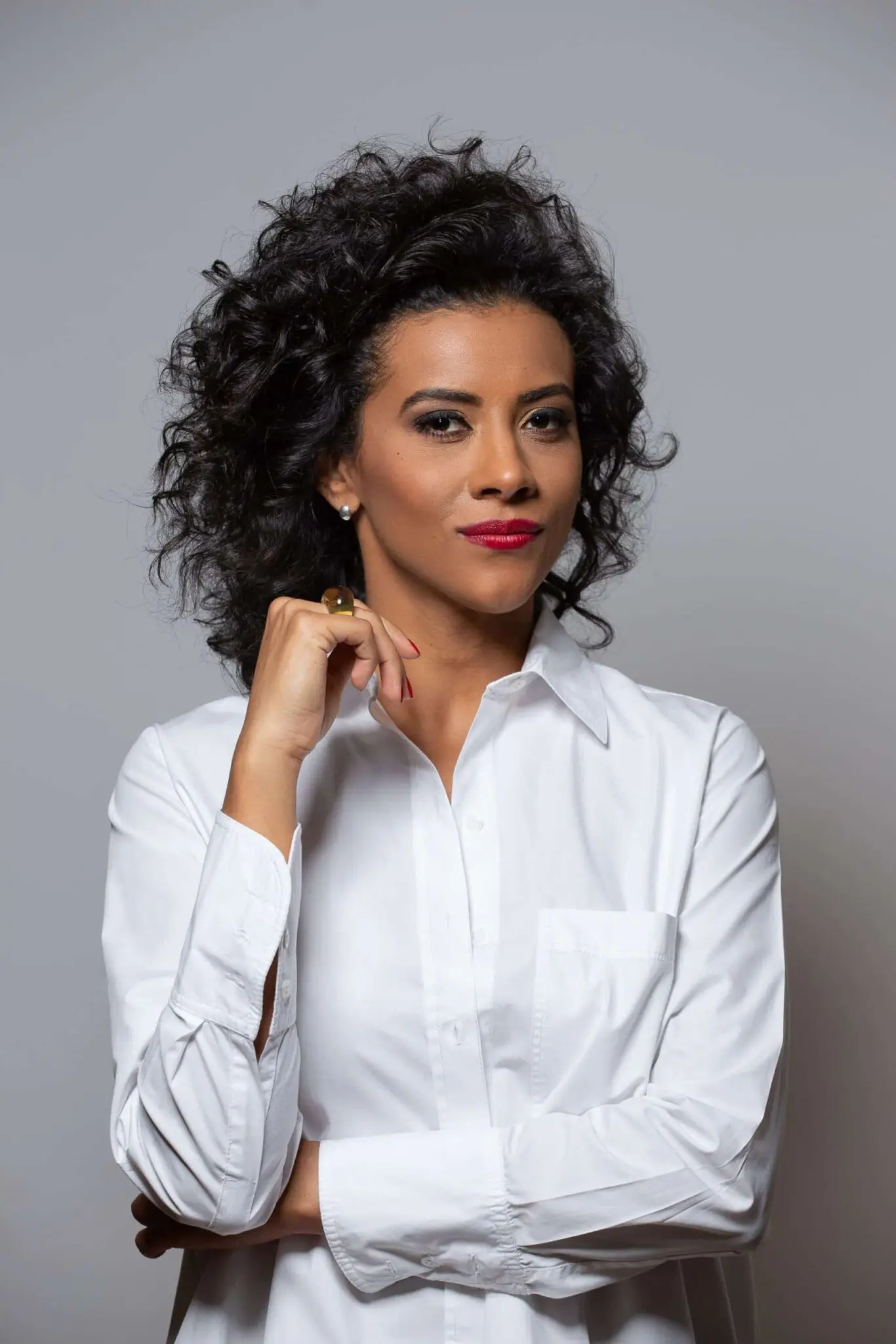 Mulher com cabelos cacheados pretos, camisa branca, braços cruzados, dedo no queixo, fundo cinza claro.