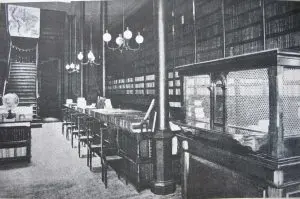 Biblioteca antiga com prateleiras de livros, escada com tapete vermelho, mesas de madeira e luminárias.
