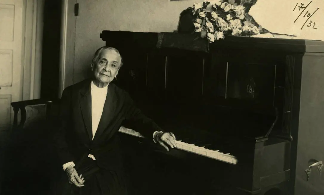 Uma pessoa em terno preto e camisa branca sentada ao piano, data "17/10/32" ao fundo.