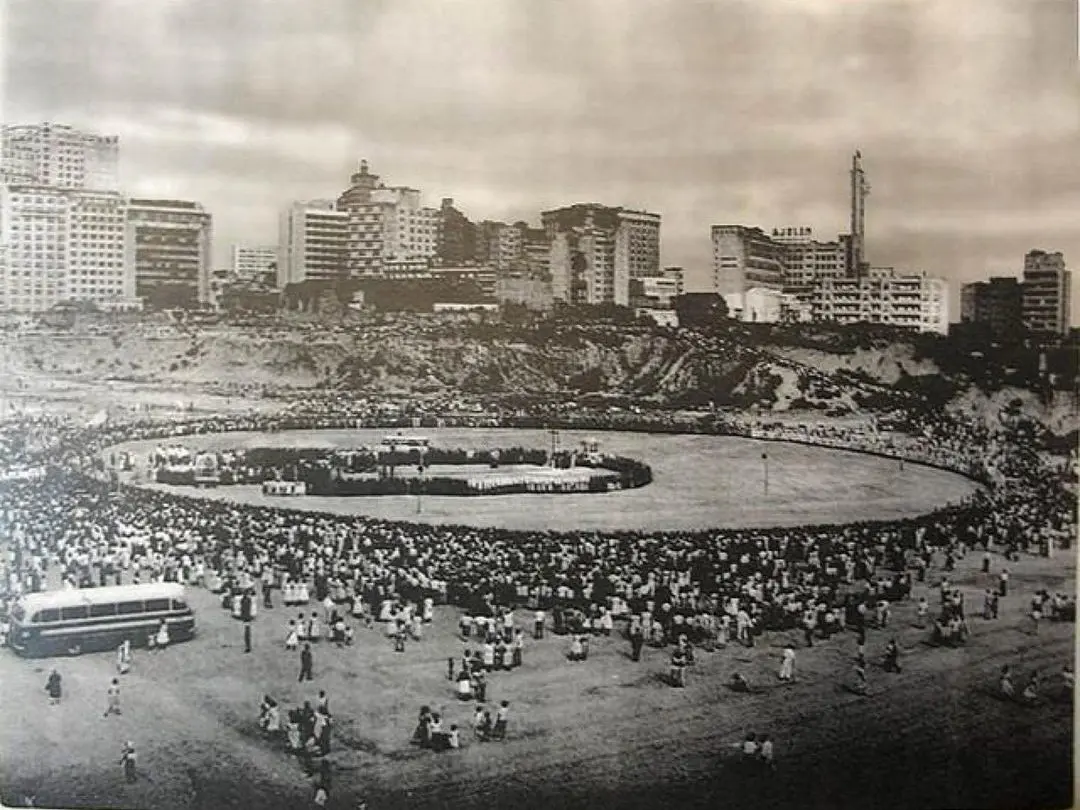 Multidão em praça, ônibus e cerimônia sob céu nublado.