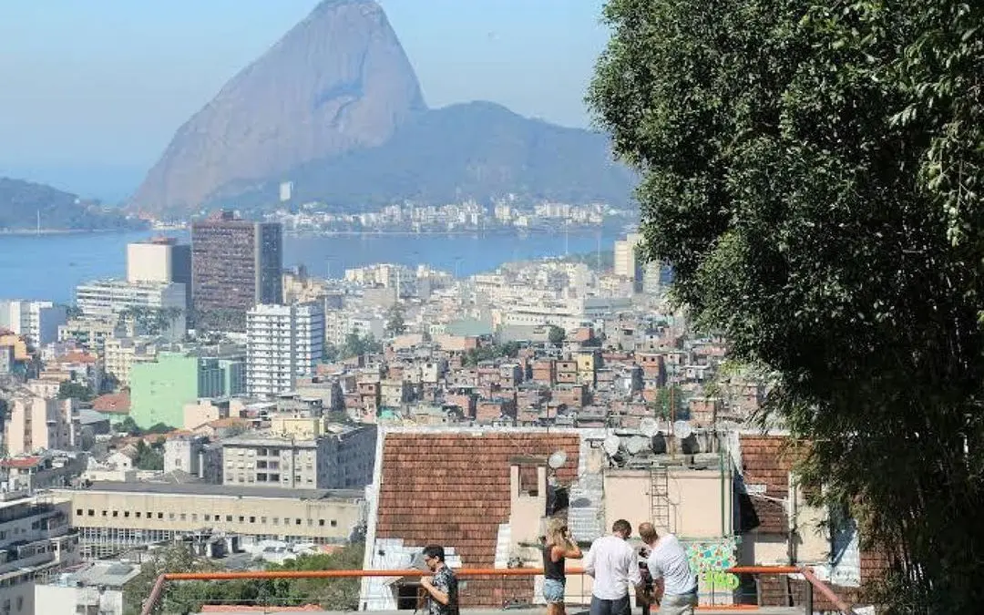 Cidade com vista para morro, edifícios variados, casas coloridas, árvore folhosa, mar e montanhas.