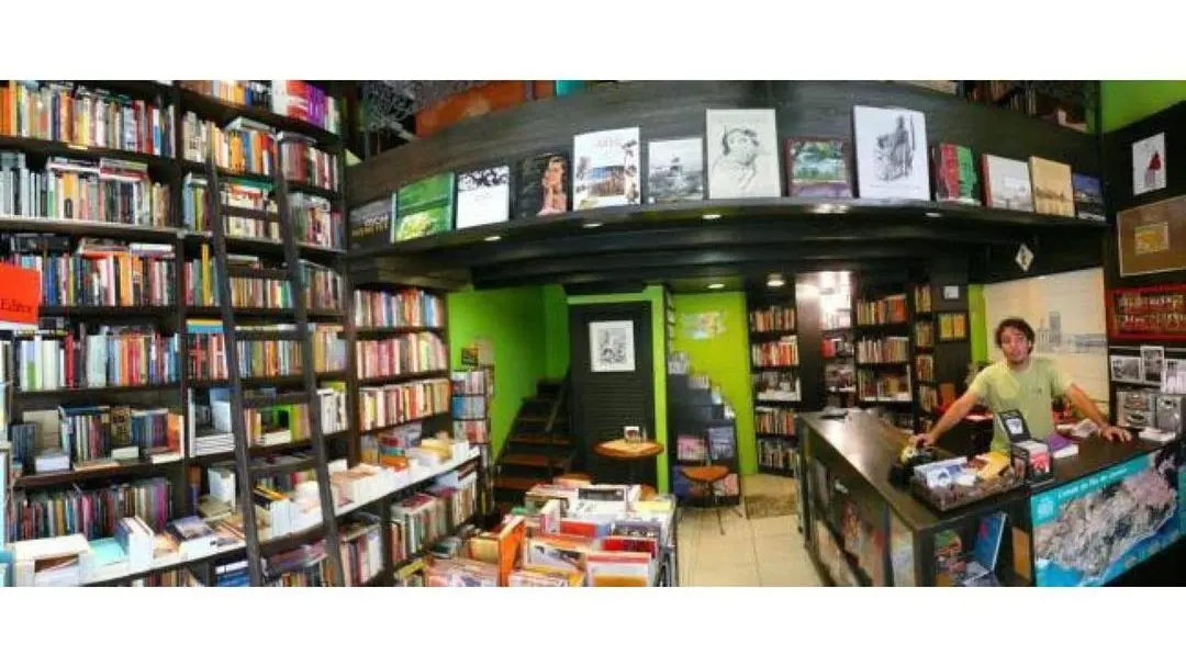 Livraria "Livraria e edições Folha Seca" com prateleiras, escadas e funcionário.