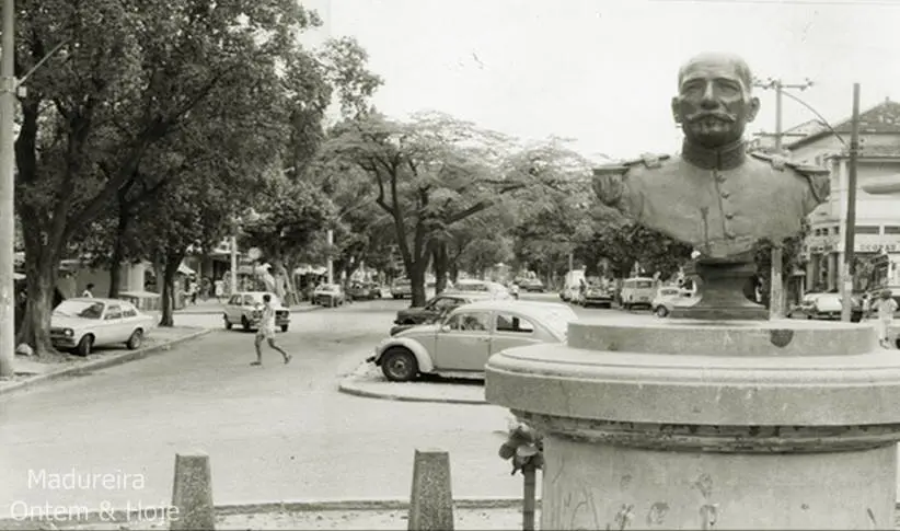 Praça com árvores verdes, carros antigos e estátua militar.