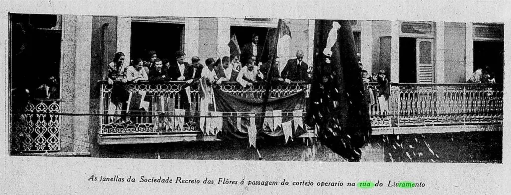 Fachada de edifício com varandas e pessoas em trajes formais, cortejo operário na rua do Livramento.