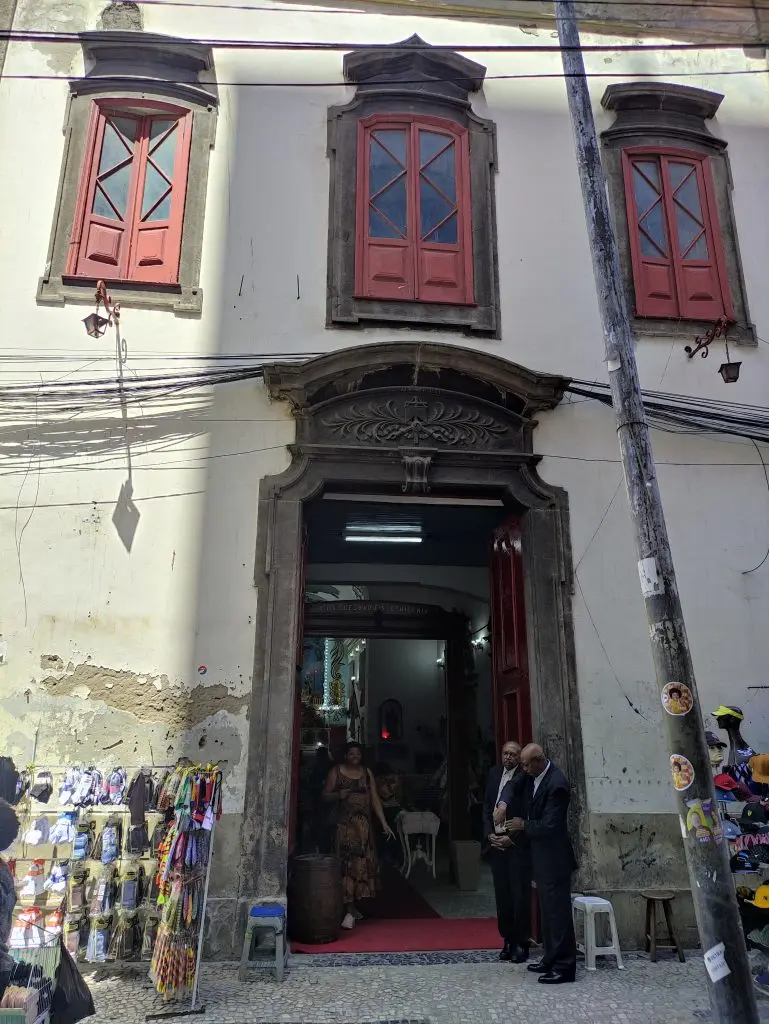Fachada colonial com entrada aberta, lojinha à esquerda e homens à direita.