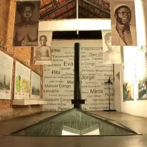 Exposição histórica com placas informativas, painel de nomes e datas, fotos de pessoas e triângulo de vidro.