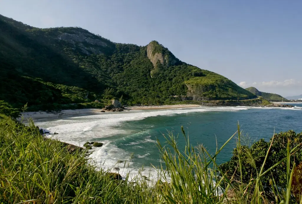 Praia com montanha ao fundo, colina verde à esquerda, penhasco central, ondas azuis, céu azul claro.