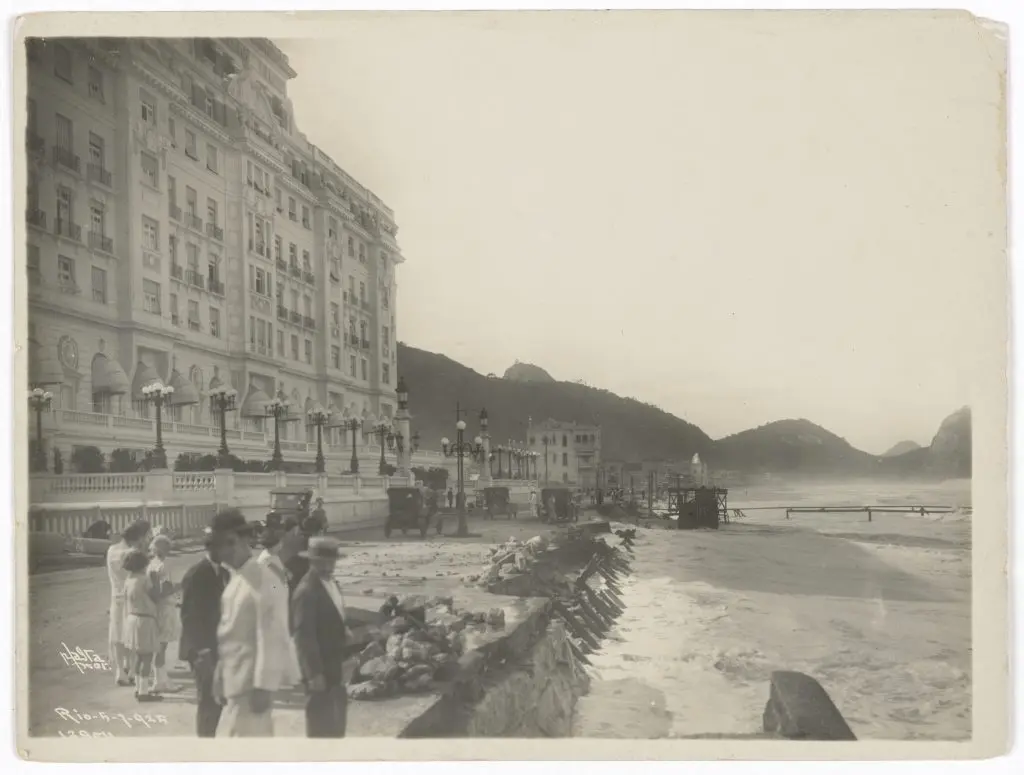 Praia antiga com hotel, calçada, muelle, ondas e montanhas.