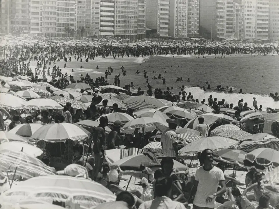 Praia lotada com guarda-sóis coloridos e pessoas na areia e na água.