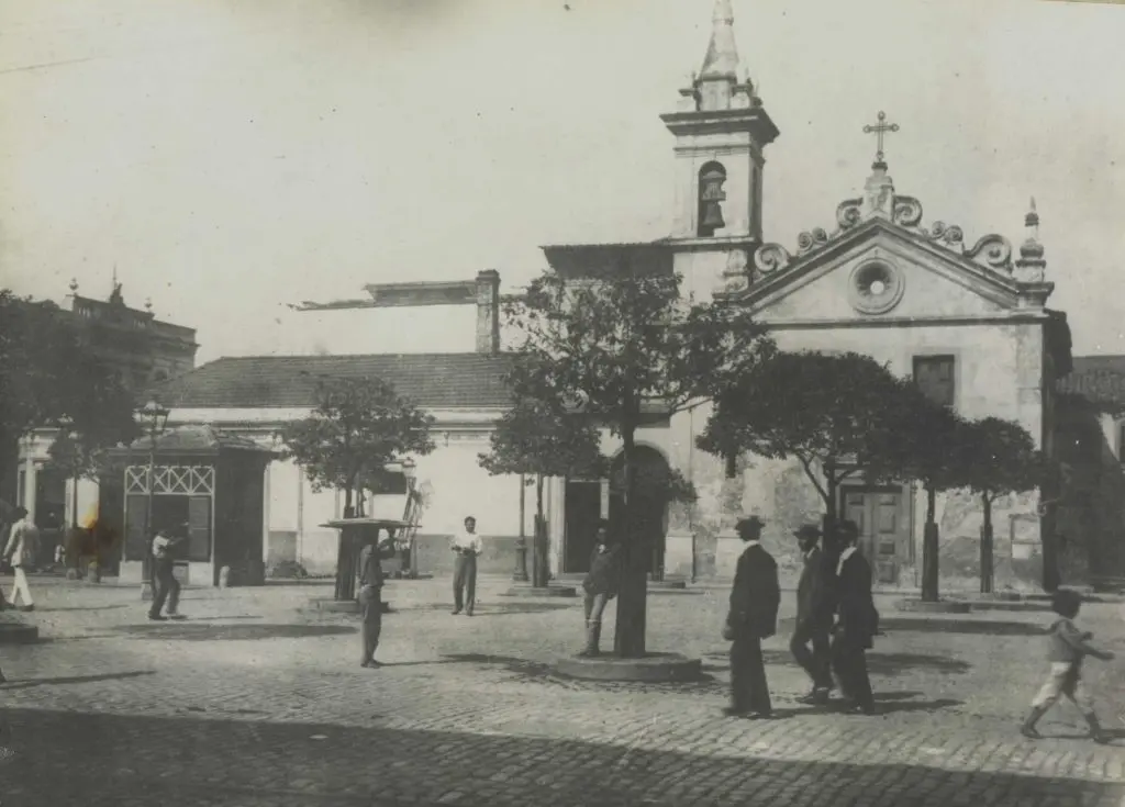Praça histórica com igreja, árvores e monumento.