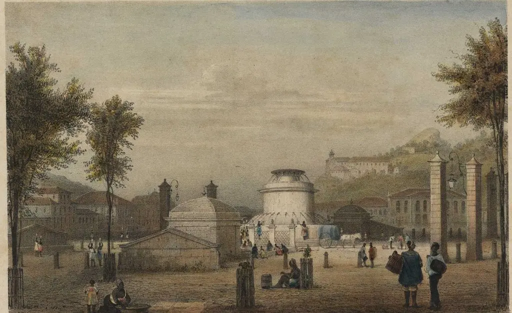 Praça histórica com árvores verdes, edifícios com cúpulas, estrutura arquitetônica e pessoas caminhando.
