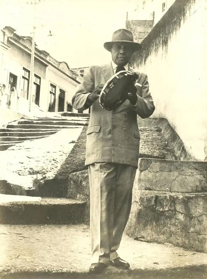 Homem em terno clássico segurando tambor em rua com construção ao fundo.