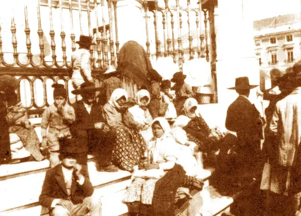 Pessoas em trajes tradicionais em banco, grade metálica ao fundo.
