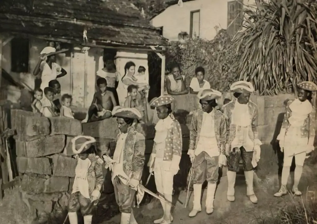 Crianças sentadas em parede de tijolos, adultos em trajes tradicionais caminham à direita, casa com porta aberta ao fundo.