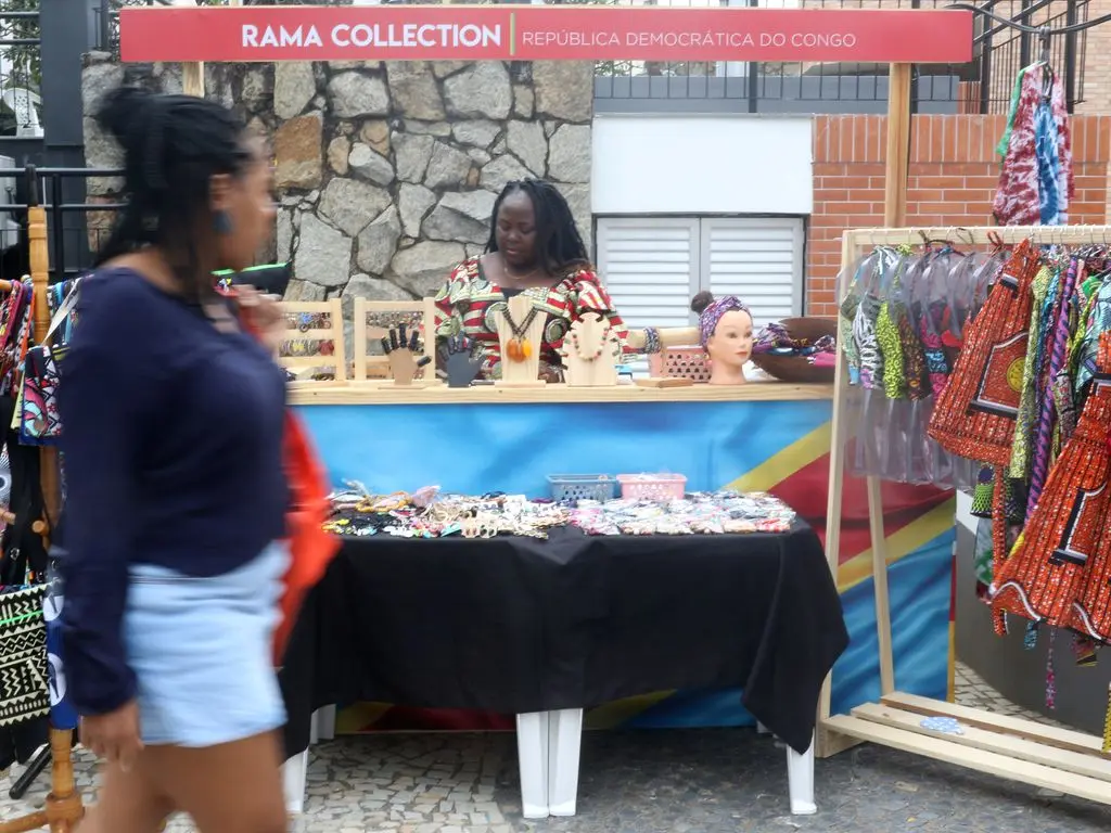 Estande comercial com mulher caminhando, joalheria e bolsas coloridas.