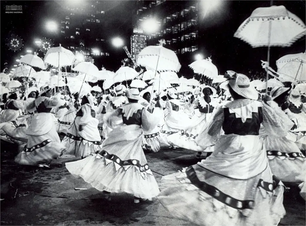 Procissão noturna com mulheres dançando, trajes tradicionais e guarda-sóis.