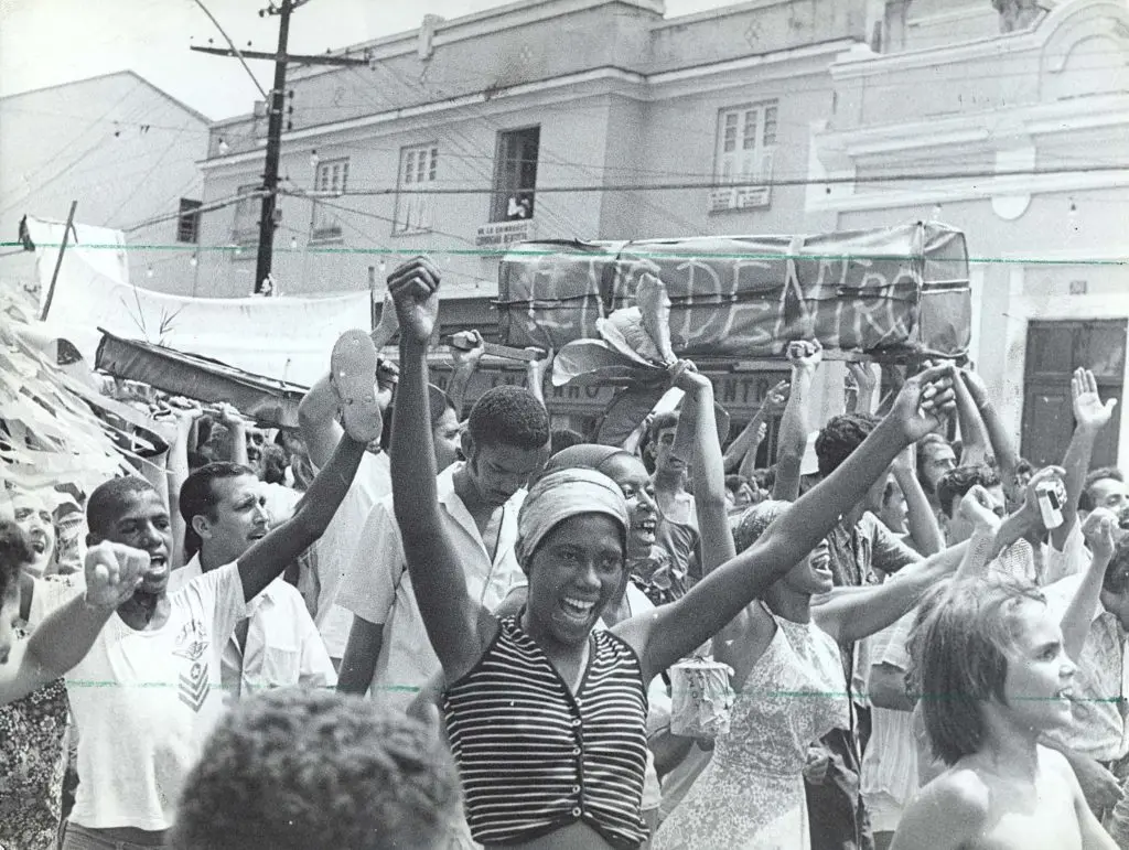 Manifestação pública em preto e branco, com multidão erguendo braços e objetos.