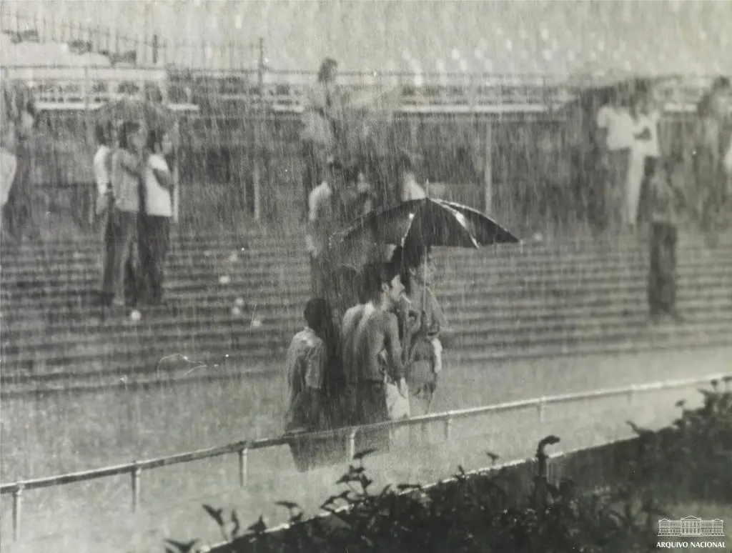 Estádio lotado durante chuva intensa, com pessoas protegidas por guarda-chuvas.